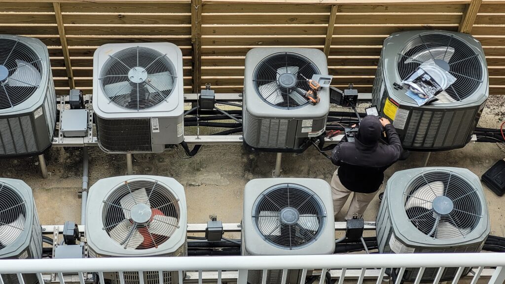 Technician servicing multiple rooftop commercial HVAC condenser units arranged in rows, performing maintenance with tools on an outdoor platform.