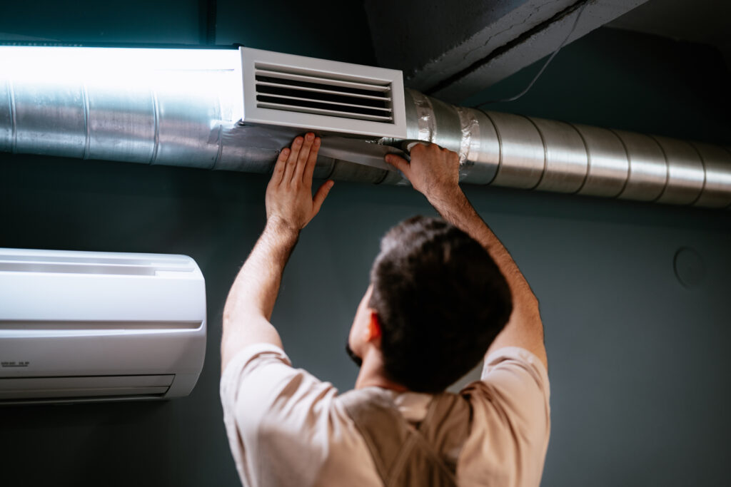 A man is focused on adjusting an air duct in a modern room. It's evening, and the indoor lighting highlights the sleek design elements of the space.