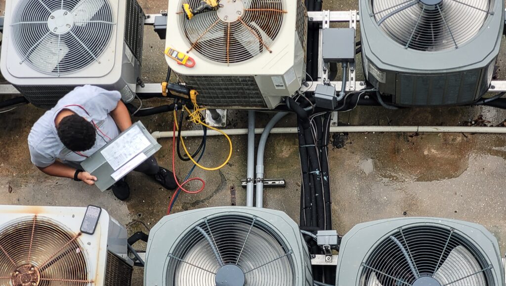 Overhead view of an HVAC technician inspecting multiple outdoor air conditioning condenser units with gauges and tools on a commercial rooftop.